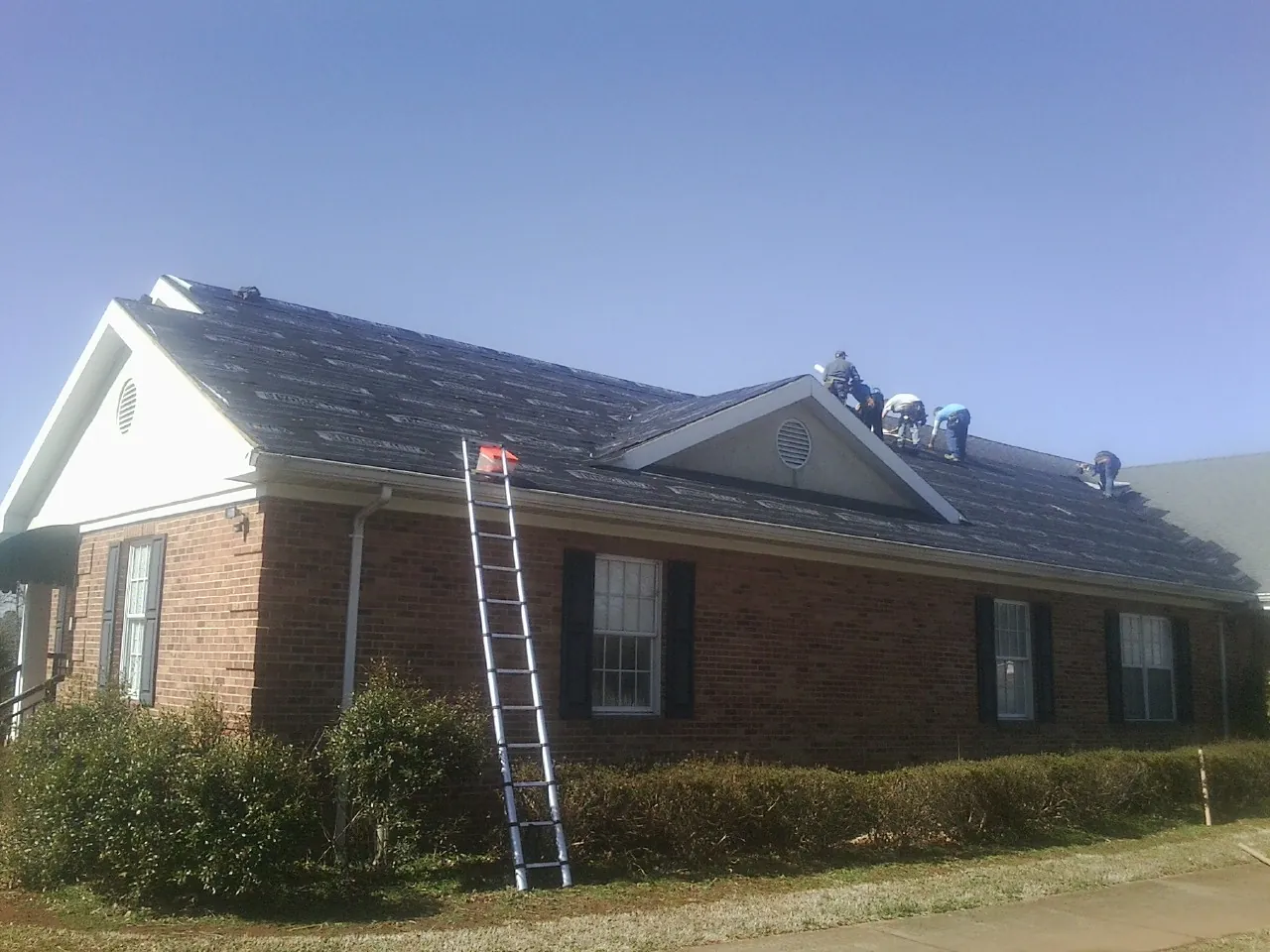 Crew tearing shingles off a brick ranch roof with a ladder set — Emergency Roof Repair work by our crew in Nettleton