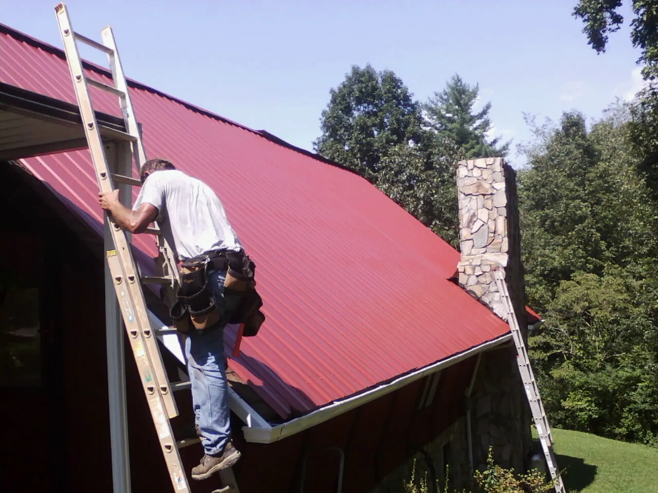 Roofer on a ladder beside a red metal roof and stone chimney — Soffit & Fascia Repair by Roofing Brothers in Nettleton